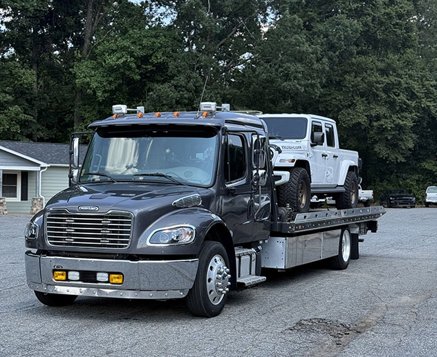 A black tow truck carrying a white Jeep Gladiator, set against a backdrop of trees and a house, parked on a gravel lot.