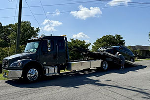 A black tow truck is loading a car on a residential street under a clear blue sky, with a person guiding the process.