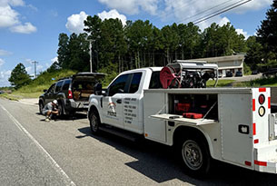 A white service truck assists a driver fixing a flat tire on the roadside, surrounded by trees and clear blue skies.