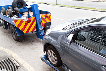A gray car is being loaded onto a blue tow truck with visible tires on the back, parked on a city street.